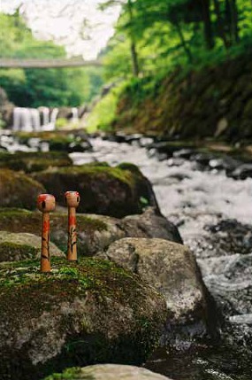 Two Kokeshi dolls on a mossy rock beside a river